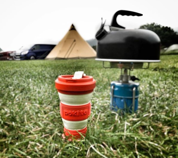 An eco-friendly red Pokito cup is being used while on a camping trip and is sitting in the grass next to a camping stove and tea kettle.