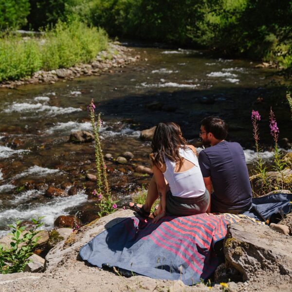 The stash mat by Rumpl is a great waterproof, stain resistant mat that can be used during your next picnic or hike; photo shows models using it to sit on a rock by the river.