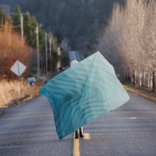 Model is walking down the street, holding her Original Puffy Blanket by Rumpl behind her, showing off the Cascade Fade Green motif.