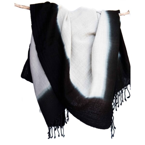 The Bloom & Give merino wool throw is hanging from a tree branch. The throw is hand dipped and dyed and has a black perimeter, black tassles, and a beige middle.