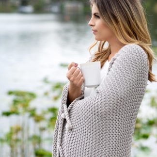 Model is shown wearing the eco-friendly Zestt Organics gray knitted throw while standing near a pond and drinking coffee.