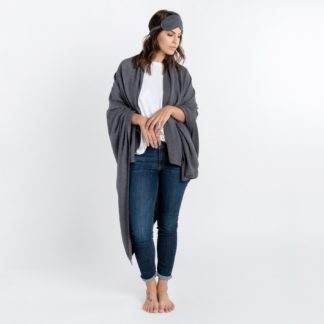 Pictures shows a model standing up, wearing the organic cotton knit throw and eye mask, in dark gray, from the Zestt Organics Cotton Dreamsoft Travel Set.
