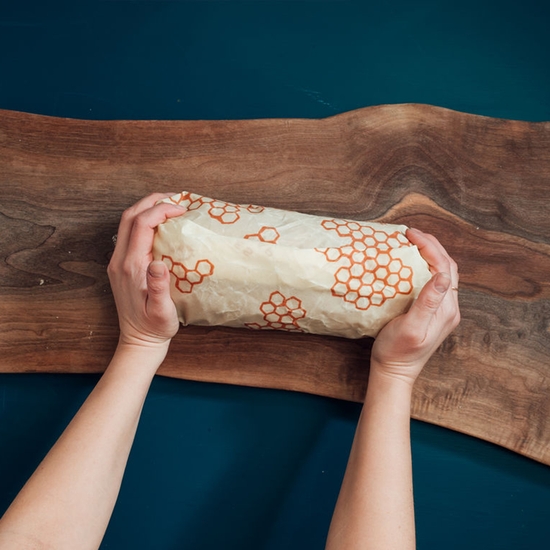 The eco-friendly Bee's Wrap brand honeycomb printed bread wrap is shown here. The model is warming the wrap with her hands so that the wrap will bend and fold around the bread.