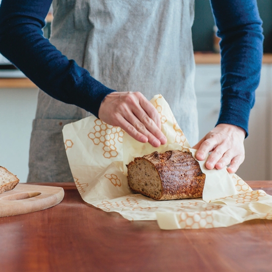 A baker is using the Bee's Wrap brand bread wrap to store a loaf of bread and is shown wrapping the bread up in the bread sized beeswax food wrap.