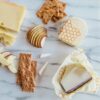 The fully biodegradable Beeswrap brand cheese wraps are shown on a granite slab, wrapped hunks of cheese, surrounded by crackers and apples.