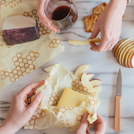 The fully biodegradable Beeswrap brand cheese wraps are shown on a granite slab, wrapped hunks of cheese, surrounded by crackers and apples.
