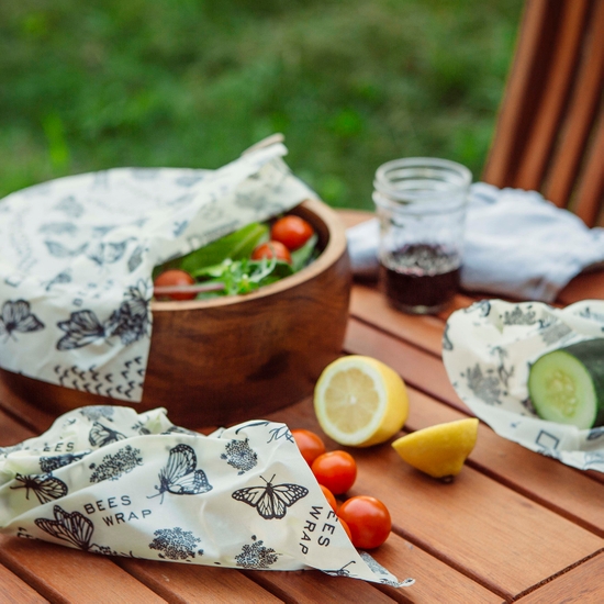 Photo shows an outdoor lunch with salad bowl and fresh fruit, covered and wrapped with Bee's Wrap branded beeswax food wraps. Shown in a fun National Geographic partnered monarch print.
