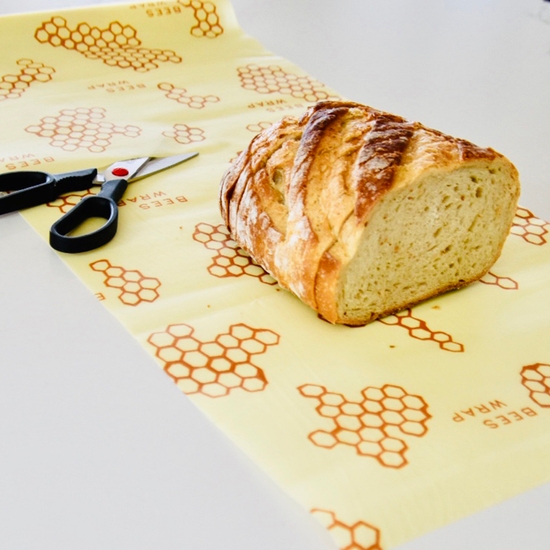 The photo shows a sheet of Bee's wrap food wrap being cut into a large size bread food wrap, with a loaf of bread sitting on top of the honeycomb wrap.