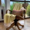 Hilana brand ultra-soft, eco friendly yellow Yalova blanket/throw is neatly folded on top of a wooden chair in someone's living room, perfectly complimented by a red patterned pillow.