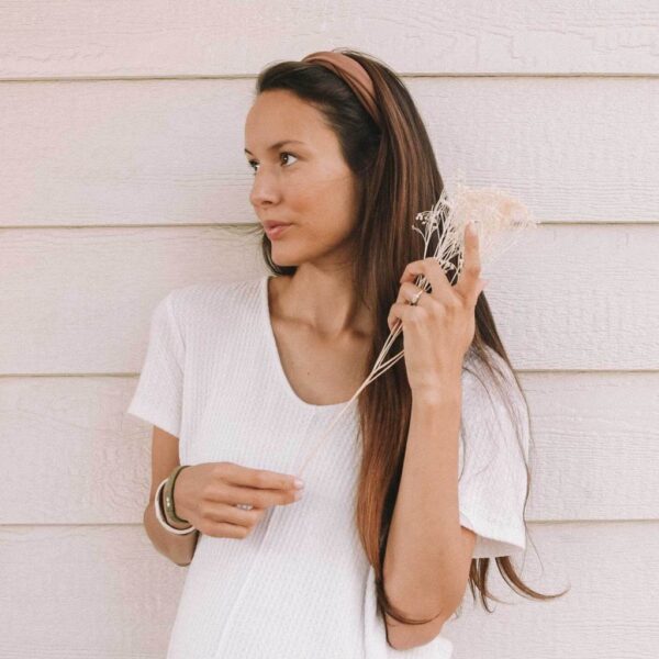 A model is holding a flower and wearing a brown eco-friendly Kooshoo brand twist headband, made with organic cotton.