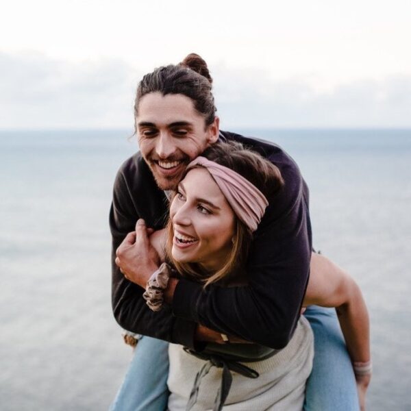 Two models are on the beach. One is wearing a Kooshoo organic cotton twist headband in pink and giving the other model a piggyback ride.