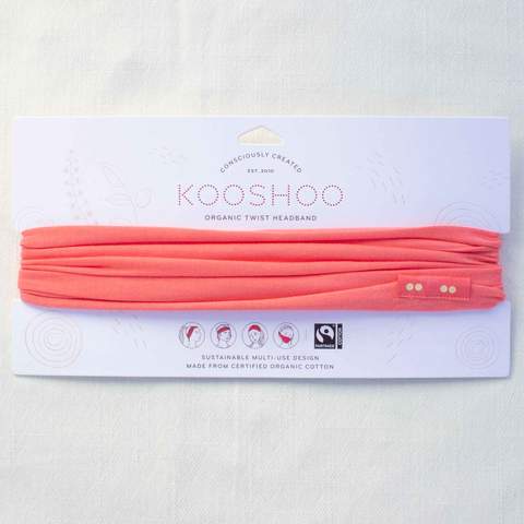 A sugar coral Kooshoo brand twist headband, made with organic cotton, is shown on display with its FSC certified compostable paper packaging.