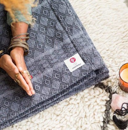 This cotton blanket from Manduka features a beautiful Peruvian pattern and is made from 87.5 percent recycled fabrics. Shown being used during a yoga practice by someone wearing gold jewelry next to a lit candle.