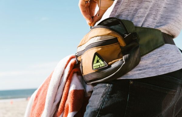 A male model on the beach, wearing his Packster hip pack from Green Guru in Earthtone colors. Hip packs are great for hiking, running, or a fun beach day!