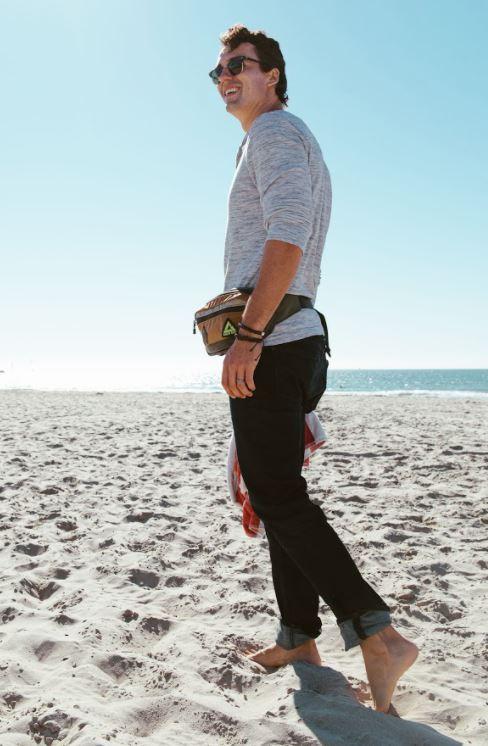 A male model on the beach, wearing his Packster hip pack from Green Guru in Earthtone colors. Hip packs are great for hiking, running, or a fun beach day!