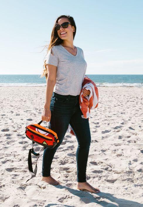 A model at the beach holding her earth-friendly Packster hip pack, by Green Guru, by the top carrying strap. Comes in fun and wild neon colors.
