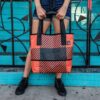 Model is wearing the Alchemy Goods one-on-a-kind sustainable billboard tote bag on her shoulder. The tote bag is made from upcycled mesh billboard fabric and comes in a variety of bright colors depending on availability.
