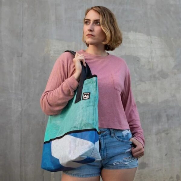 Model is wearing the Alchemy Goods one-on-a-kind sustainable billboard tote bag on her shoulder. The tote bag is made from upcycled mesh billboard fabric and comes in a variety of bright colors.