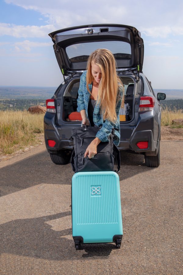 Model has taken the Meridian luggage by Sherpani out of the back of her car; you can see the teal blue color of the luggage with black wheels and accents.