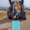 Model has taken the Meridian luggage by Sherpani out of the back of her car; you can see the teal blue color of the luggage with black wheels and accents.