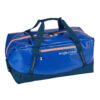 The sustainable migrate duffel bag by Eagle Creek, shown here in mesa blue, is a 90 liter duffel bag with blue fabric, orange zippers, and navy straps that is made from durable and weather-resistant recycled materials.