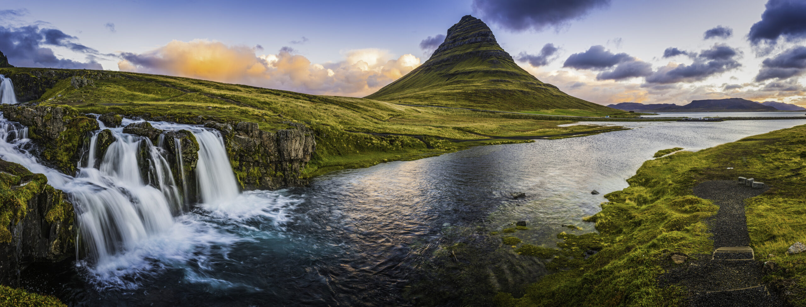 The clear blue waters of Kirkjufellfoss waterfall tumbling down the mountainside to the ocean fjord beside Grundarfjordur beneath the iconic peak of Kirkjufell framed by the warm glow of sunrise over the Snaefellsnes peninsula in the far east of Iceland for sustainable travel.