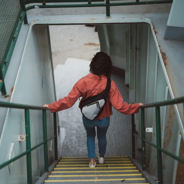 This image shows a woman walking down stairs with the Esprit AT Shoulder Sling bag over her back. She's wearing the sterling color.