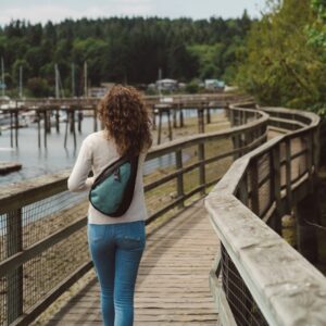 The Esprit AT Shoulder Sling Bag is seen here, in teal, by someone wearing the pack on their back as they go for a walk. It has anti-theft technology and is made from 100% recycled materials.
