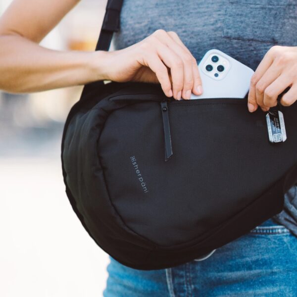 This image shows somebody putting their cell phone into the front pocket of the Esprit AT Shoulder Sling Bag. They are wearing the bag on their front side, giving easy access to the front zipper pouch that fits a cell phone. The bag is shown here in Carbon.