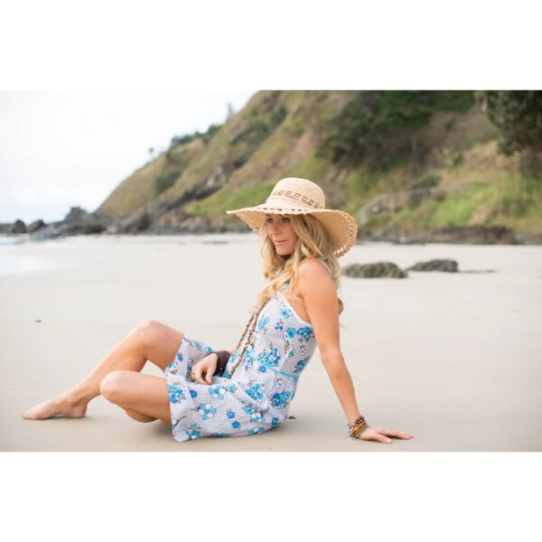 This shows a woman sitting on the beach wearing the natural colored Amy Summer Raffia Hat. The wide brim offers sun protection and the vented crown allows for cool air flow. This hat only comes in one color and one size.